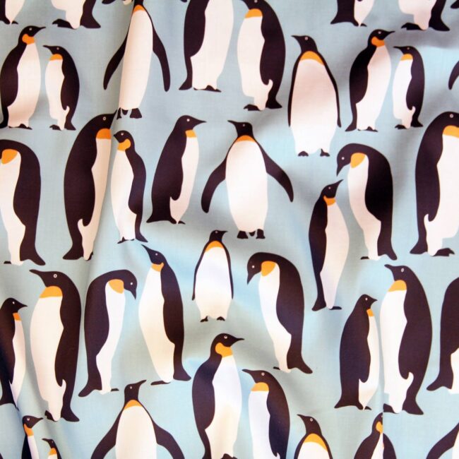 Close-up of a textile pattern featuring a densely repeating print of black, white, and yellow Emperor penguins on a pale blue background.