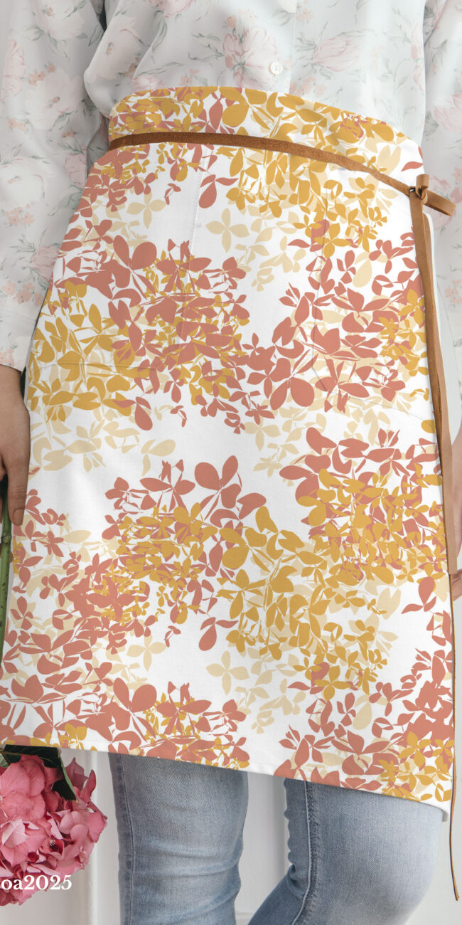 Woman wearing a brown leather-tie half apron with an abstract, repeating pattern of orange, coral, and yellow autumn leaves, holding a pink hydrangea flower.