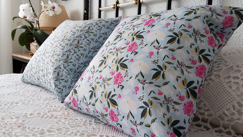 Close-up of two large square throw pillows on a bed with white crochet lace bedding, featuring a light blue fabric with a detailed pattern of white, pink, and green flowers and leaves.