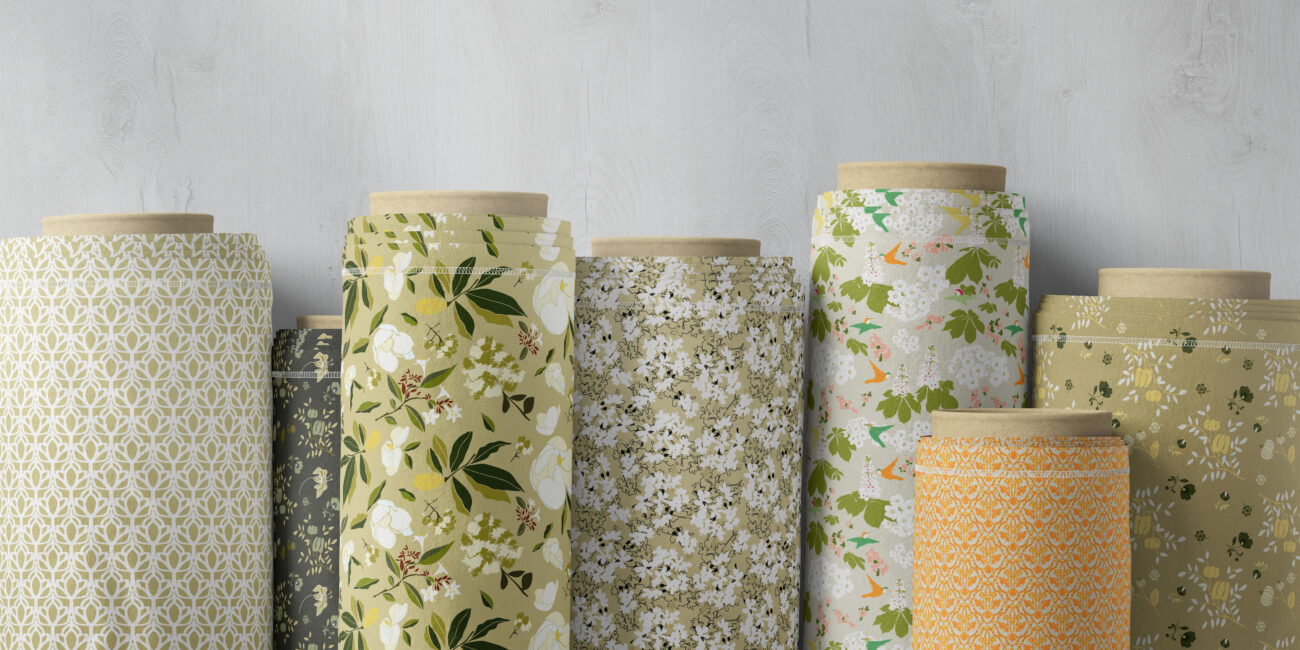Seven rolled fabric bolts showcasing natural and organic botanical print patterns in shades of white, sage green, and earthy yellow.