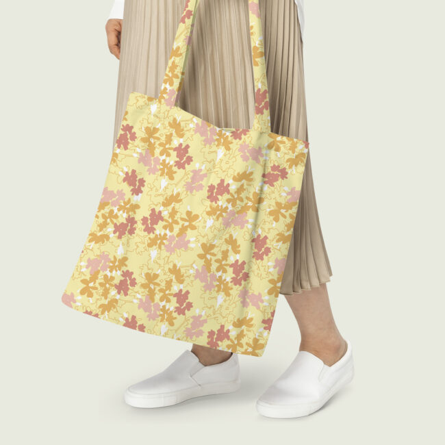 Person holding a yellow canvas tote bag with a repeating pattern of stylized pink and orange flowers, against a neutral background.